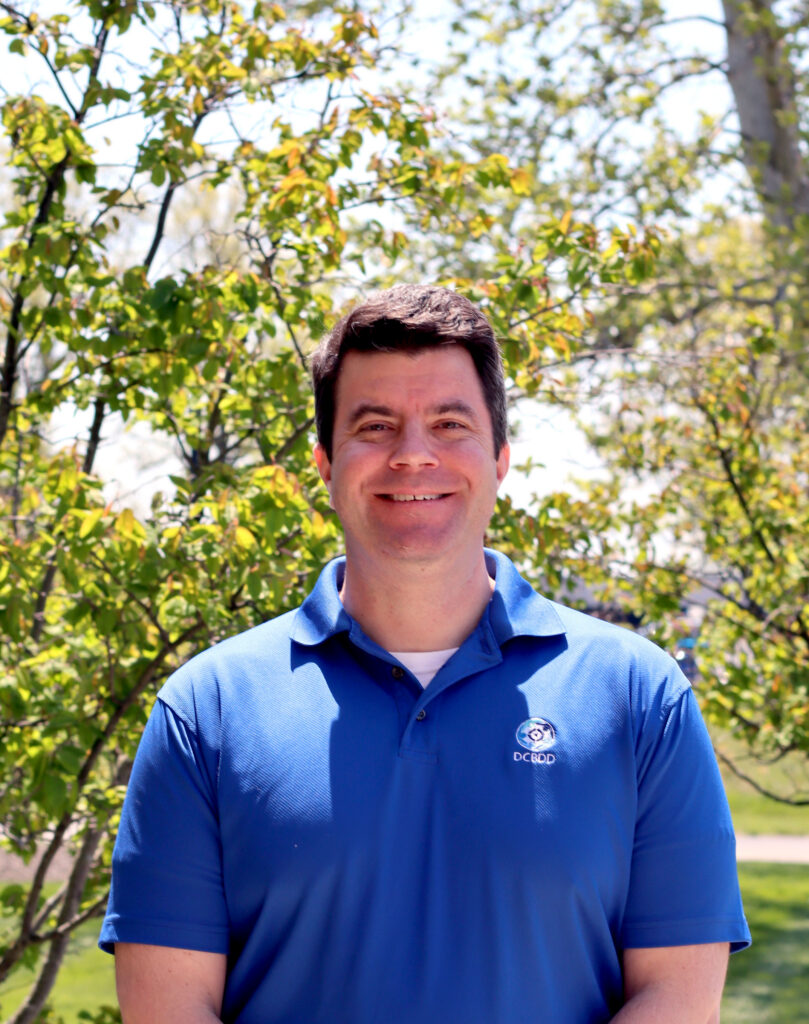 A person smiling outdoors in front of green trees, wearing a blue collared shirt with a logo.