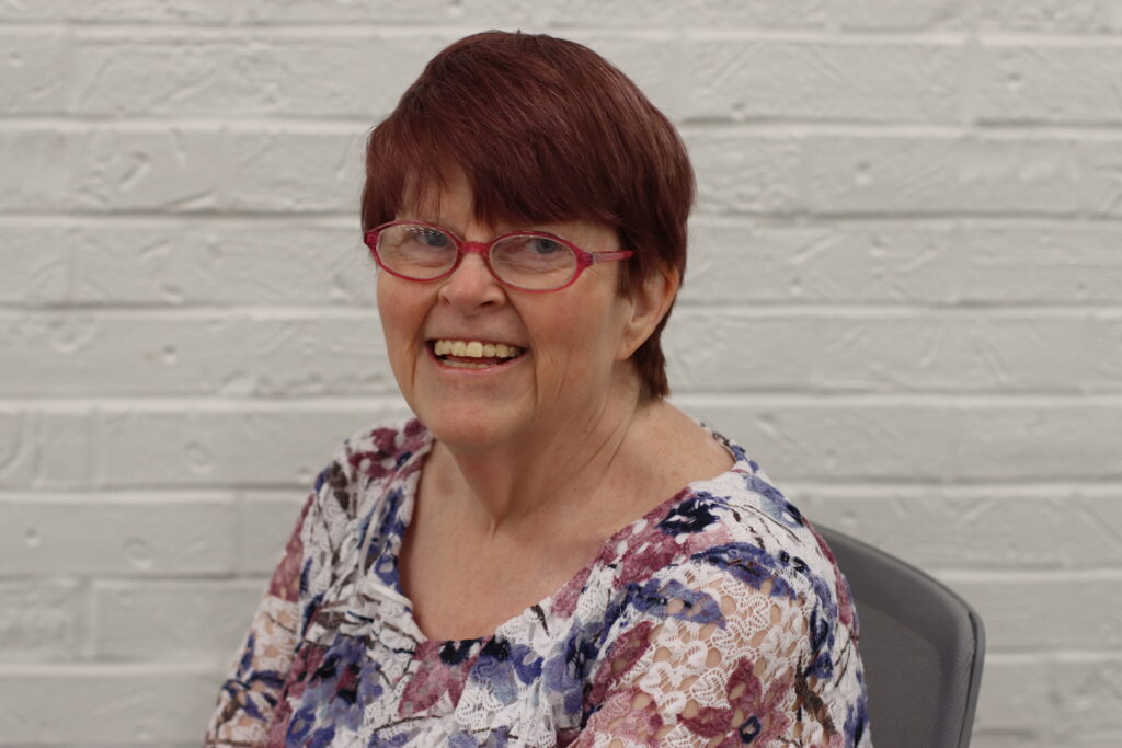 A person with short reddish-brown hair and red glasses smiling in front of a white brick wall.