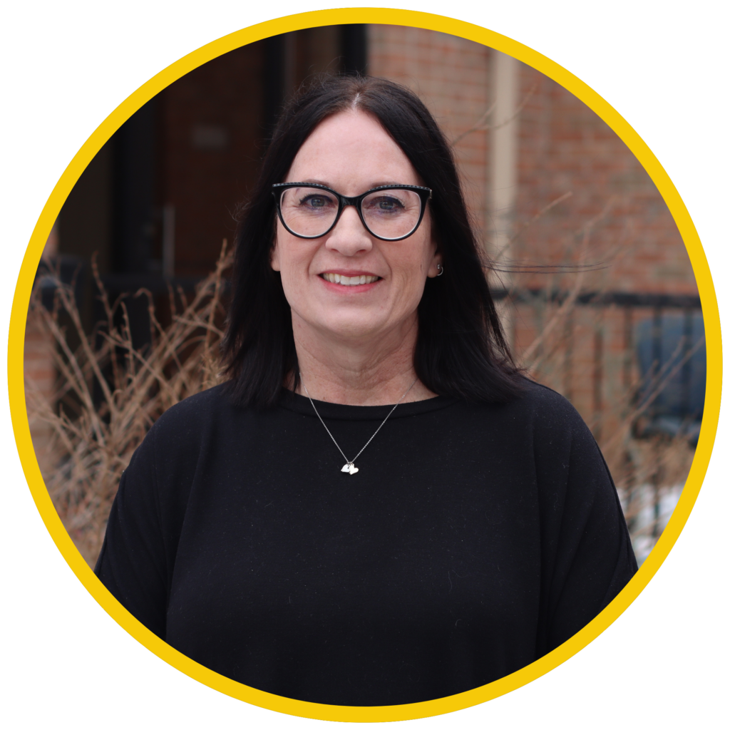 A woman with dark shoulder-length hair and black glasses smiles at the camera while standing outdoors in front of a brick building. The photo is framed in a circular yellow border.