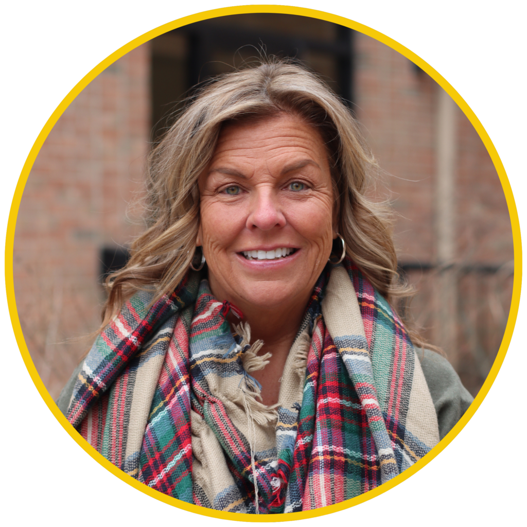 Susan smiles at the camera in an outdoor portrait, wearing a plaid scarf, hoop earrings, and a green jacket, framed inside a circular yellow border.