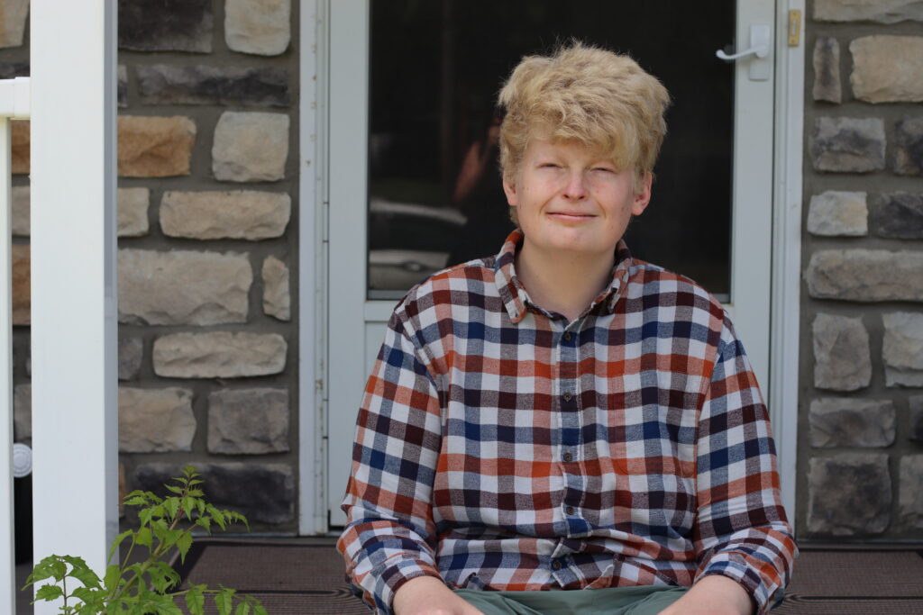 Ian sits on a wooden porch wearing a plaid flannel shirt, in front of a stone wall and a white door.
