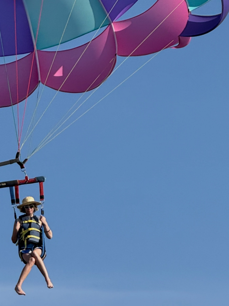 Lauren in a life vest parasailing beneath a colorful purple, teal, and magenta canopy against a blue sky.