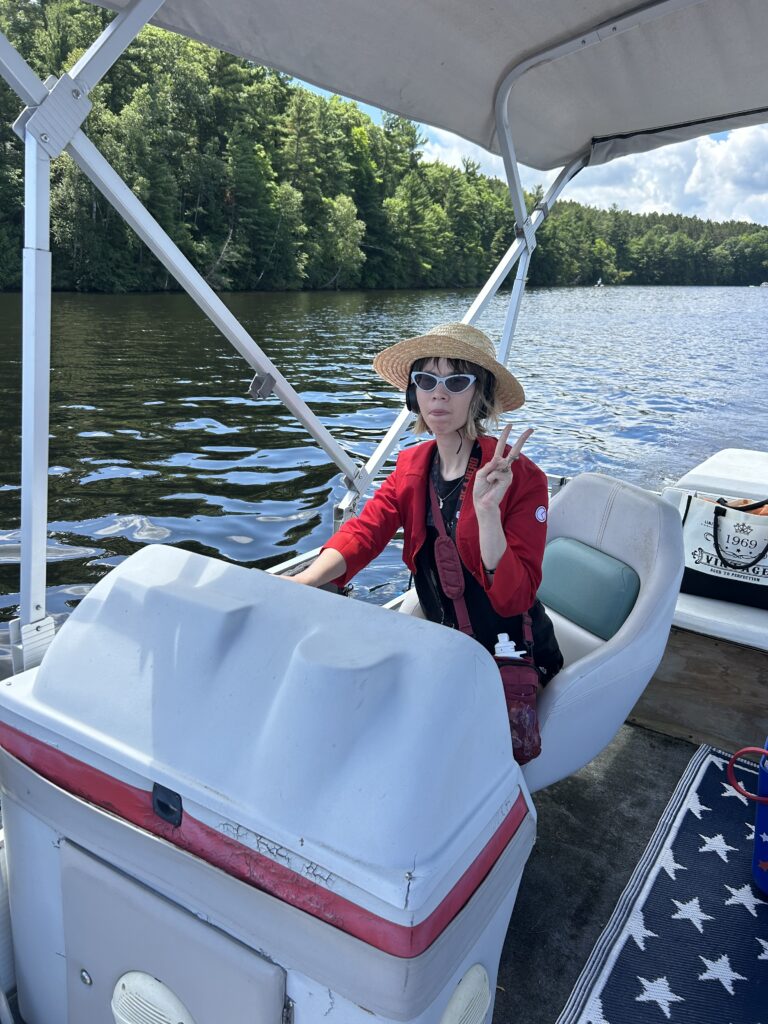Lauren wearing a straw hat and sunglasses gives a peace sign while steering a covered boat on a tree-lined lake.