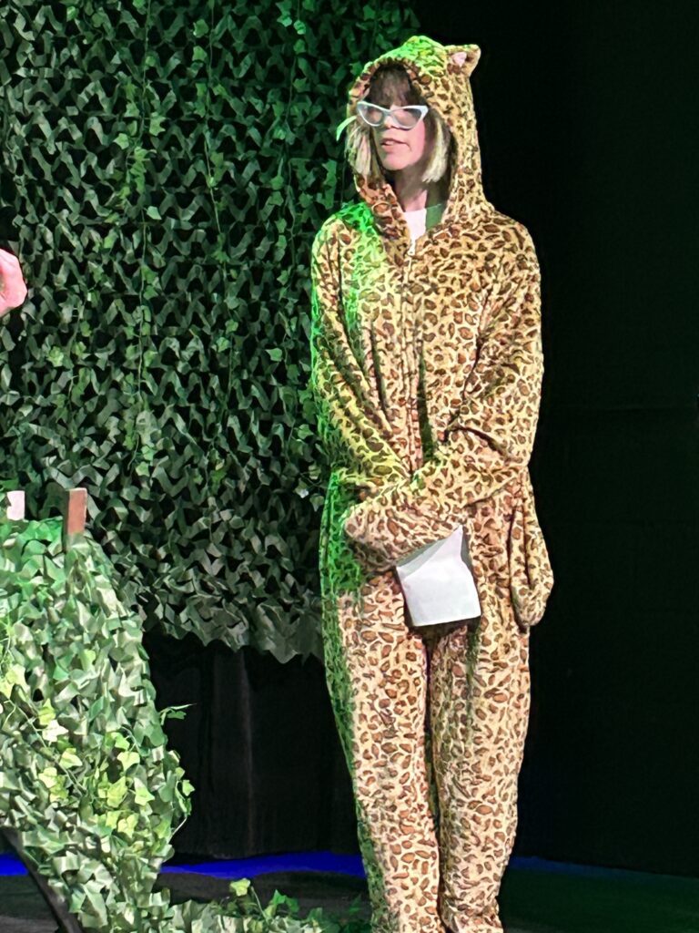 Lauren in a leopard-print animal costume and white glasses standing on a foliage-covered stage.