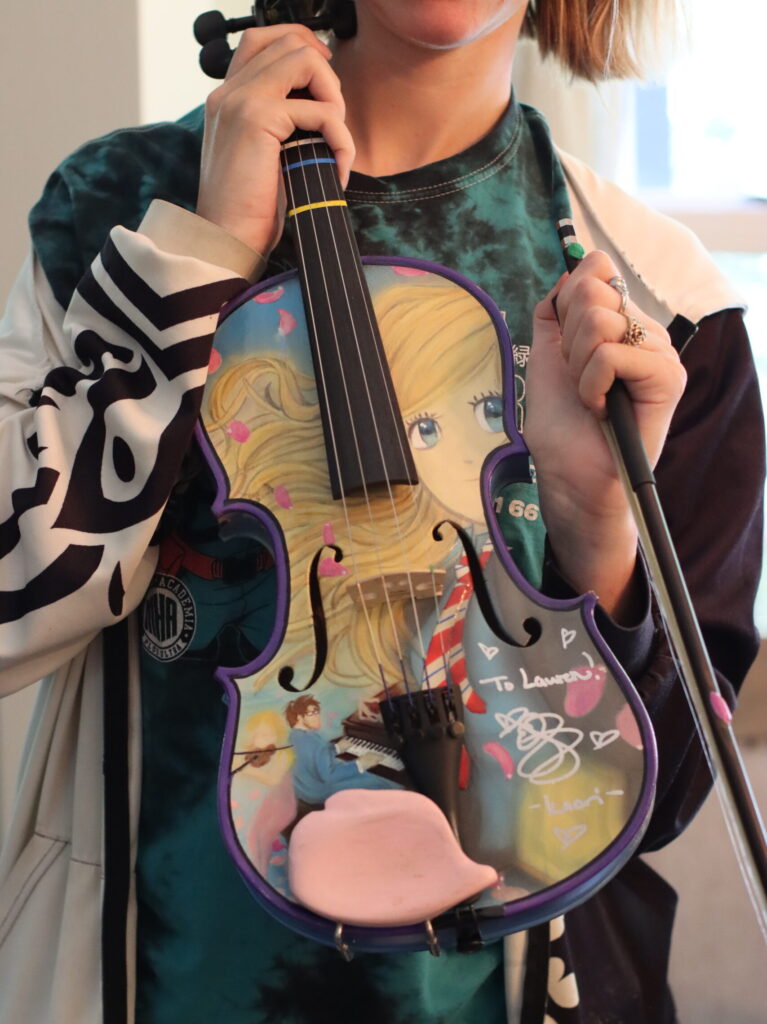 Lauren holding a colorful, hand-painted violin with purple trim, character illustrations, and handwritten messages.