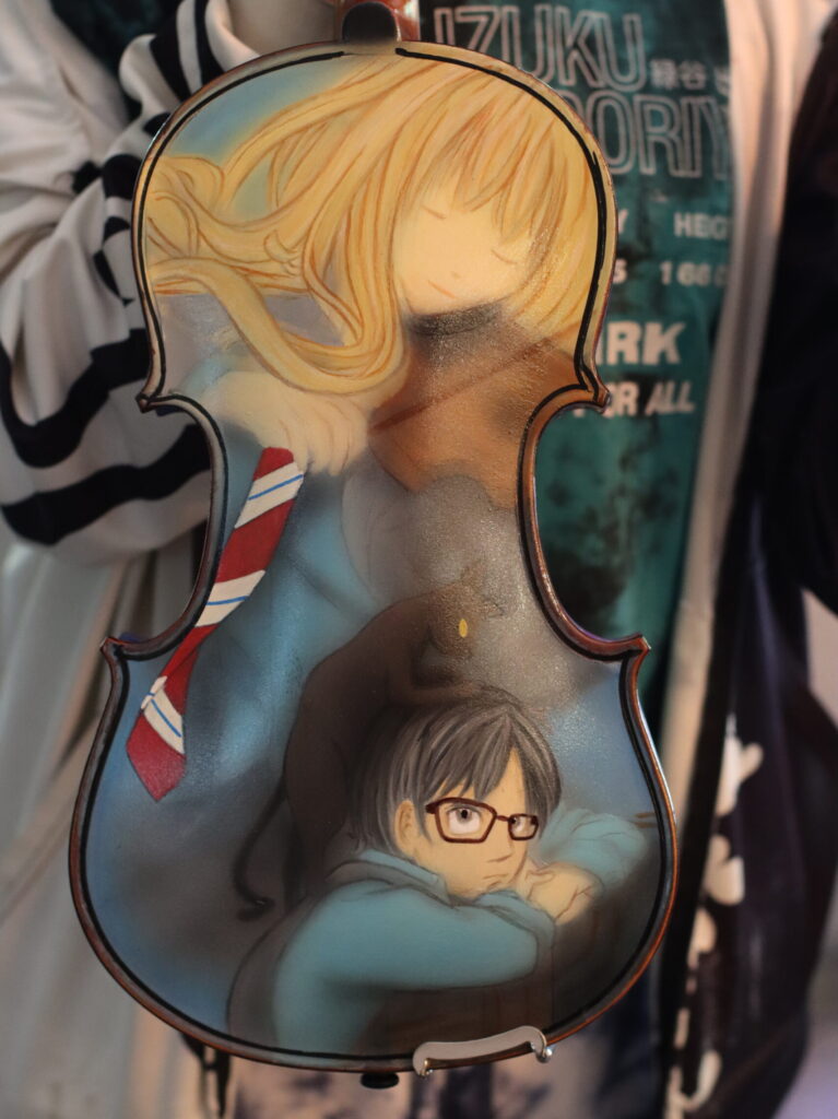 A vertically oriented, violin-shaped decorative object fills the center of the image. It has a dark outline, a glossy painted surface, curved sides, and small metal fittings along the top and bottom. The artwork shows a large, pale-skinned figure with long golden-blond hair across the upper half, and a smaller dark-haired figure wearing rectangular glasses and a blue garment in the lower half. A red, white, and blue striped tie-like design appears along the left side. The object is photographed against a dim, crowded background with people and clothing; a green shirt with partially obscured lettering is visible behind it.