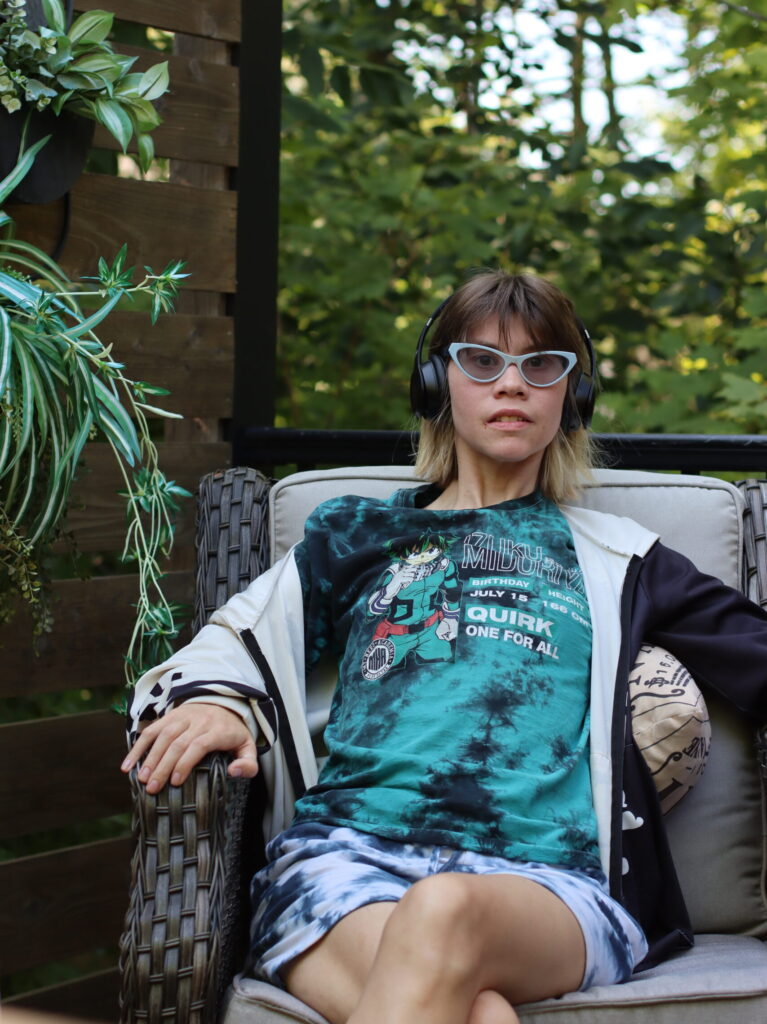 Lauren wearing white cat-eye glasses and headphones seated in a patio chair among plants and trees.