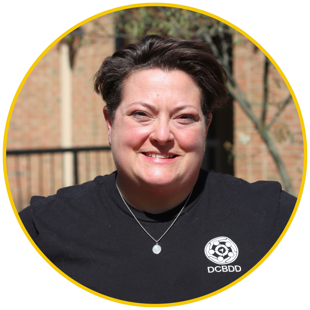 Smiling adult wearing a black DCBDD shirt and silver pendant, photographed outdoors within a yellow circular border.