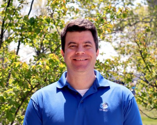A person smiling outdoors in front of green trees, wearing a blue collared shirt with a logo.