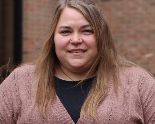 A person with light-colored hair wearing a textured pink cardigan over a black top is standing outdoors in front of a brick building.