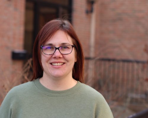 A person with medium-length hair wearing a light green sweatshirt is standing outdoors in front of a brick building.