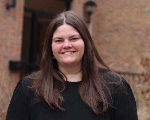 A person with long, straight hair wearing a black top is standing outdoors in front of a brick building.
